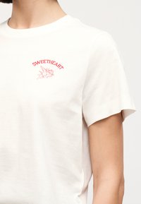 Άσπρο κοντομάνικο t-shirt με κόκκινη επιγραφή "SWEETHEART" πάνω από έναν μικρό κόκκινο Έρωτα που κρατά τόξο και βέλος στην αριστερή πλευρά του στήθους.