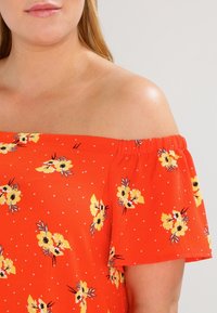 Rotes Off-Shoulder-Oberteil mit weißen Punkten, das ein florales Muster in Gelb und Schwarz aufweist. Strukturierter Stoff mit elastischem Ausschnitt.