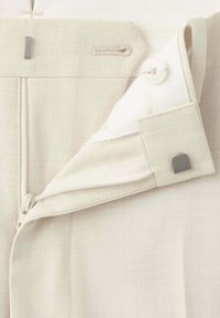 Ceinture de pantalon beige avec zip partiellement ouvert, bouton et fermeture à crochet métallique sur tissu clair.