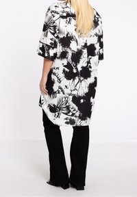 Vestido camisero de estampado floral en blanco y negro con mangas cortas, dobladillo asimétrico y textura sedosa, combinado con pantalones acampanados negros.