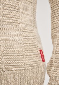 Beżowy sweter dziany z fakturowanymi wzorami. Po boku znajduje się czerwona metka z logo. Materiał wydaje się miękki i ciepły.