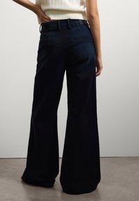 Hoge taille donkere denim wijde pijpen broek met gestikte achterzakken en een gladde textuur, gecombineerd met een witte geribbelde top.