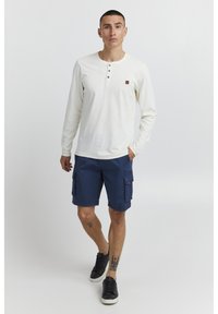 Biela dlhá tričko s dlhými rukávmi typu henley s tromi gombíkmi, spárované s námorníckymi cargo šortkami so side vreckami. Čierne tenisky dotvárajú vzhľad.
