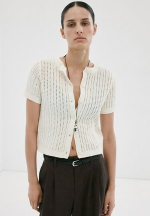 Žena oblečená v krátkym rukávom bielom pletenom zapínacom kardigáne a čiernych nohaviciach s vysokým pásom, so záveskom píšťalky na čiernom retiazku.