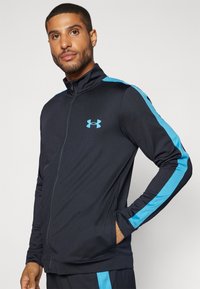 Veste de sport noire avec un col haut, ornée de rayures bleues le long des épaules et des bras. Le matériau est lisse et extensible.