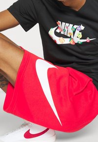 Osoba oblečená v černém tričku Nike s barevnými ovocnými logy, červených šortkách Nike a bílých teniskách se červeným logem Nike, sedící s pokrčenou nohou.