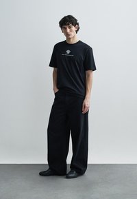 Musta puuvillainen T-paita, jossa on lyhyet hihat ja rintamuksessa graafinen kuvio, yhdistetty väljästi istuviin mustiin housuihin ja mustiin kenkiin.