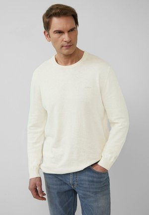 Lichtbeige lange mouw trui met een ronde hals en geribbelde manchetten, voorzien van een klein, gouden logo-detail op de borst, gedragen met blauwe jeans.