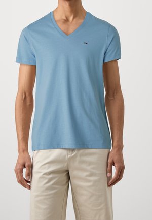 Mężczyzna w jasnoniebieskim T-shircie z dekoltem w kształcie litery V i krótkim rękawem oraz beżowych spodniach, stojący na jednolitym tle.