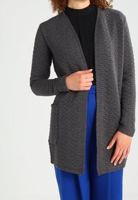 Szary pikowany kardigan z dekoltem w serek, długimi rękawami i kieszeniami z przodu, w połączeniu z niebieskimi spodniami. Wzór z fakturą dodaje szczegółów.