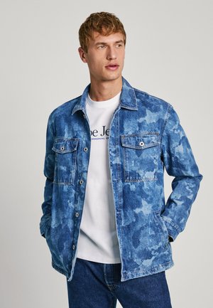Veste en denim bleu clair avec un motif camouflage, deux poches poitrine et une fermeture à boutons. Portée par-dessus un t-shirt blanc et un jean bleu.