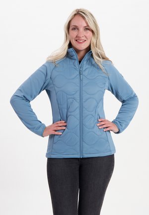 Lichtblauwe softshell jas met een ritssluiting aan de voorkant, voorzien van een golfpatroon textuur en elastische manchetten. Gecombineerd met donkergrijze broek.