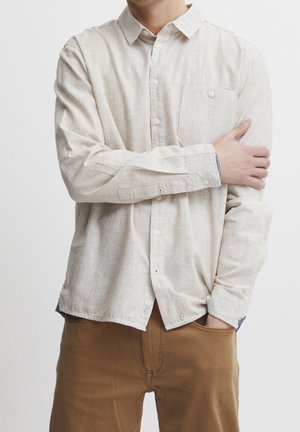 Homme portant une chemise beige clair boutonnée et un pantalon marron, une main dans la poche et l'autre tenant le bras opposé.