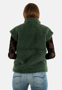 Gilet in pile verde fuzzy senza maniche con colletto alto e finitura testurizzata, abbinato a una camicia nera trasparente a maniche lunghe e jeans chiari.