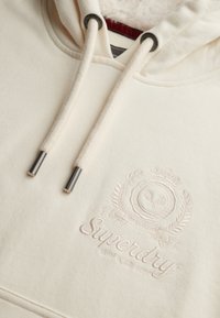 Sudadera color crema con capucha texturizada, cordones con puntas metálicas y un logo en relieve que presenta una corona y elementos decorativos.