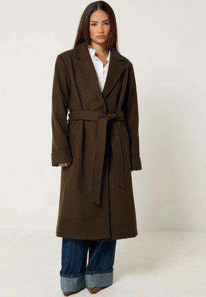Femme portant un long manteau marron ceinturé par-dessus une chemise blanche et un jean large bleu retroussé, debout sur un fond clair uni.