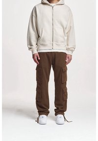 Krémová mikina na zip s předními kapsami, která je zkombinována s hnědými cargo kalhotami s několika postranními kapsami. Celý outfit doplňují bílé tenisky.
