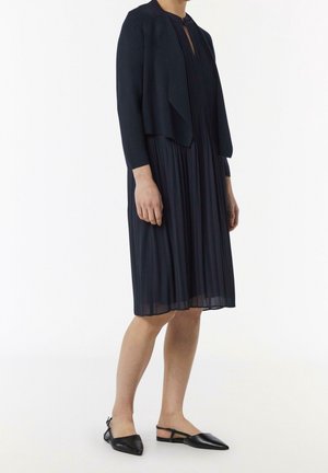 Marineblaues, plissiertes Kleid mit rundem Ausschnitt, kombiniert mit einer taillierten marineblauen Strickjacke. Schwarze spitze Ballerinas vervollständigen das Outfit.