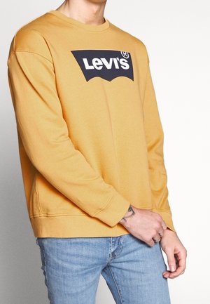 Person trägt ein senfgelbes Levi's Sweatshirt und hellblaue Jeans, mit einem sichtbaren Handgelenk-Tattoo an der rechten Hand.