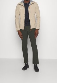 Un homme se tient vêtu d'une veste à capuche beige, d'une chemise noire, de pantalons vert foncé, de chaussettes noires et de chaussures de randonnée noires devant un fond gris uni.