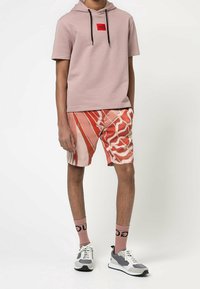 Uomo che indossa una felpa a maniche corte di colore rosa chiaro con lacci neri, pantaloni corti grafici rossi, calze rosa abbinate e sneaker grigie e bianche.