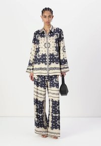 Navy and cream patterned silk pajama set with a collared button-up shirt and wide-leg pants. Model holds a small black handbag.