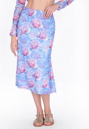 Vrouw draagt een midi-rok met blauw-roze bloemenpatroon en een bijpassend langarmige top, gecombineerd met gouden sandalen met bandjes en platte zolen, staat met één hand op haar heup.