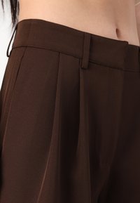 Brune skreddersydde bukser laget av glatt stoff med høy midje, plisserte detaljer i front og beltehemper for justerbar passform.