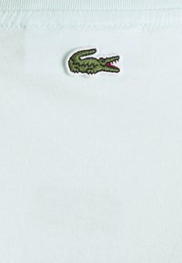 Chemise en coton bleu clair avec un logo de crocodile brodé vert. Le tissu a une texture lisse et un col côtelé.