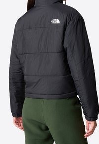 Sort pufferjakke med kviltet design, høj krave og elastiske manchetter. Logo på den øverste ryg. Båret over grønne fleecebukser.