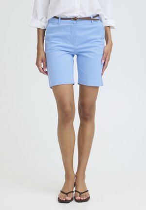 Kobieta w jasnoniebieskich szortach do kolan z brązowym paskiem, białe podwinięte rękawy koszuli i brązowe plecione sandały.