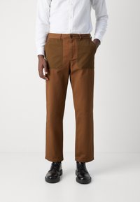 Brown cotton trousers with dual front pockets and a straight-cut design, featuring a button closure. Paired with black shoes.