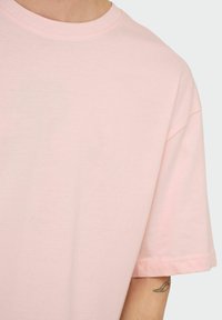 Person wearing a light pink crew neck T-shirt showing upper torso and part of a tattooed arm against a plain background.