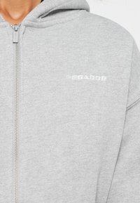 Nahaufnahme eines grauen Reißverschluss-Hoodies mit Metallreißverschluss und kleinem weißen "PEGADOR"-Logo auf der Brust.