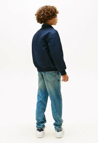 Barn med krøllet hår iført en marineblå jakke, lyseblå jeans og hvide sneakers, som står sidelæns på en hvid baggrund.