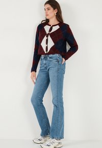 Cropped cardigan with a maroon, navy, and white argyle pattern, featuring round buttons. Paired with light blue flared jeans and sneakers.