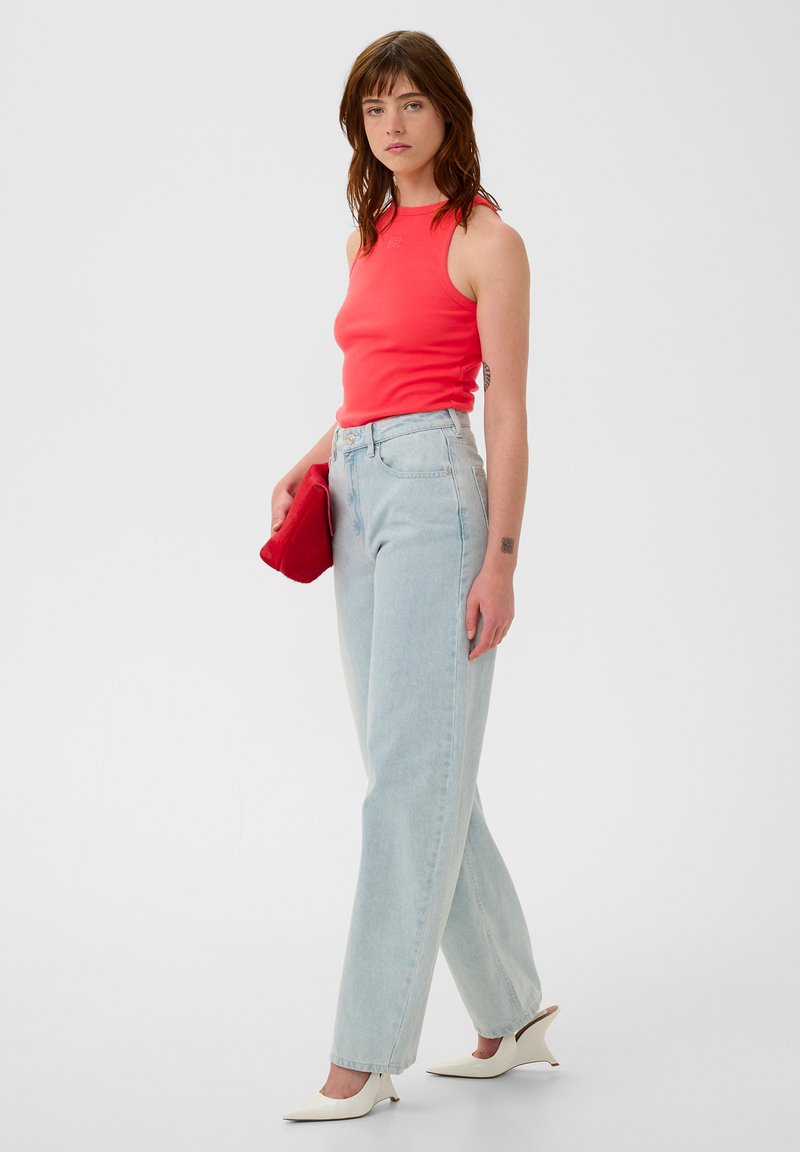 Red sleeveless top, high-waisted light blue jeans, holding a red clutch. Pointed white heeled shoes. Simple, fitted design with minimal detail.