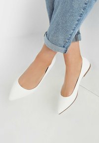 Białe płaskie buty z ostrym noskiem wykonane z gładkiego materiału, charakteryzujące się minimalistycznym wzornictwem i płaską podeszwą, noszone z podwiniętymi dżinsami.