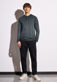 Langärmliges Henley-Shirt aus dunkelgrünem Stoff mit drei Knöpfen am Hals. Kombiniert mit schwarzen Hosen und cremefarbenen Sneakers.