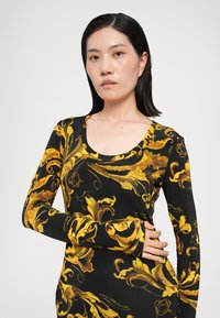 Svart långärmad klänning med ett guld barockmönster, tillverkad av stretchtyg med rundad halsringning och figursydd design.