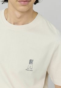 T-shirt en coton beige avec un petit graphique d'items alimentaires empilés et le texte "Hungry Games" en dessous, doté d'un col rond classique.