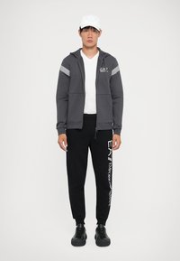 Sweatshirt cinza com fecho, mangas às riscas, t-shirt branca, calças de moletom pretas com logotipo em texto e sapatilhas pretas. Modelo masculino em pé.