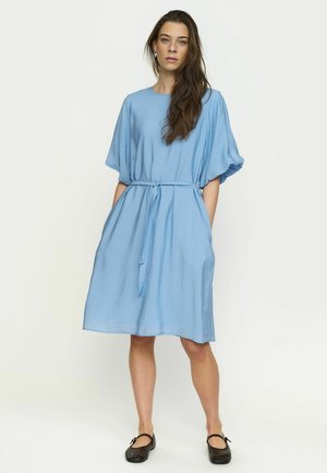 Pruunide pikkade juustega naine, kes kannab lahtist helesinist põlvpikkust kleiti vöököitega ja musti madalaid kingi, seisab valge tausta ees.