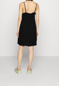 Černé šaty bez rukávů s volánkovým výstřihem, lehký materiál, volný střih a detail na zádech. Spárováno se zelenými platformovými botami.