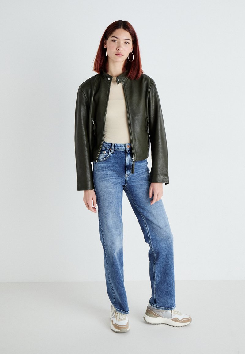 Chaqueta de cuero verde con cuello alzado, camiseta beige, jeans azules y zapatillas beige. El modelo está de pie sobre un fondo neutro.