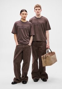 Brune bomuld T-shirts med præget "TOMMY JEANS" logo, kombineret med matchende løstsiddende bukser. En tanfarvet duffeltaske med brune detaljer holdes.