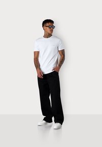 Weißes T-Shirt mit kurzen Ärmeln und einem kleinen rosa Logo auf der Brust, kombiniert mit schwarzen weiten Hosen und weißen Sneakers. Flache Textur, lässige Passform.