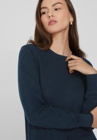 Suéter de punto azul marino con mangas largas y cuello redondo; tejido texturizado visible; el modelo toca suavemente el cuello con una mano.