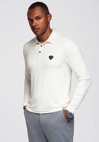 Polo a maniche lunghe color crema con colletto, tre bottoni neri e un logo triangolare sul petto; abbinato a pantaloni grigi a righe.