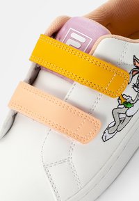 Sneaker bianco con cinturini in velcro gialli e pesca, linguetta in tessuto rosa e design di personaggio ricamato sul lato.